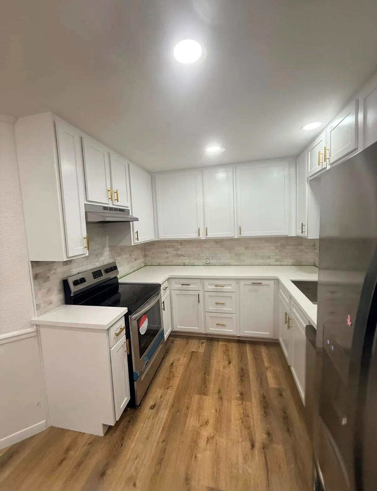 Finished galley kitchen: white shaker cabinets with brass pulls, quartz-look counters, stacked stone backsplash and new wood-look plank floor.