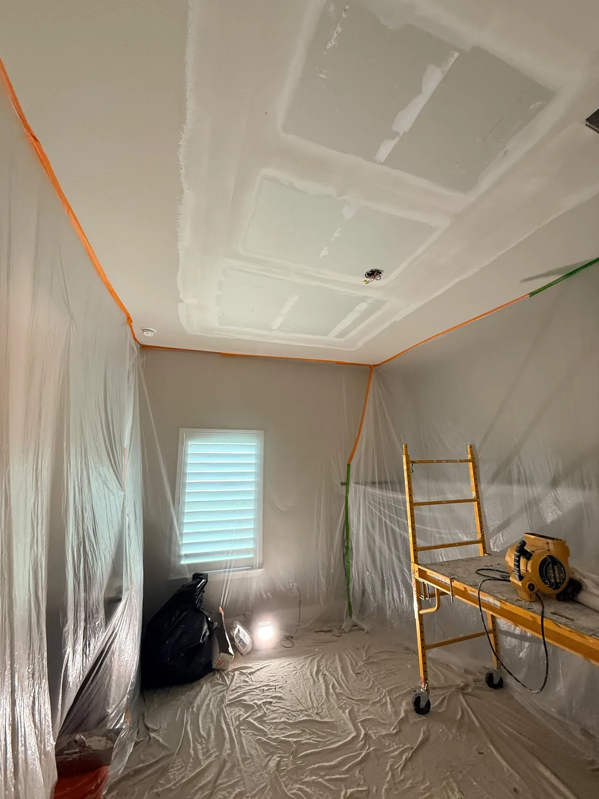 Ceiling drywall floated and taped with the walls fully plasticked off and the floor covered — scaffold and fan in place for drying.